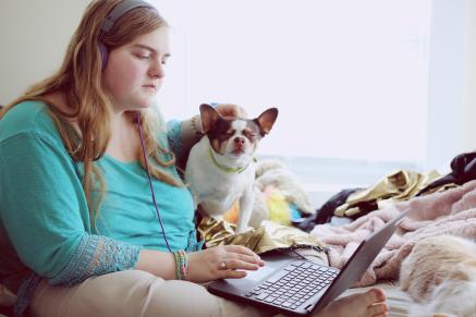 A teen girl with a dog sits reading a laptop.