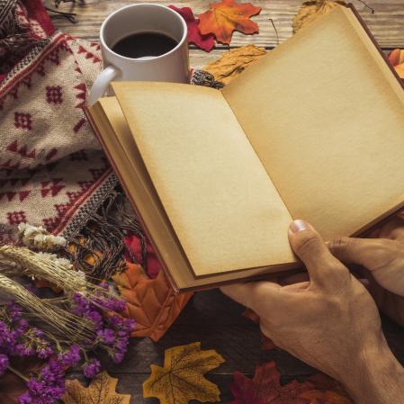Hands holding a book on a wooden table, surrounded by red and brown fall leaves and a cup of tea