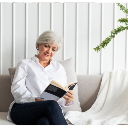 A senior woman sits on a couch, reading a book.