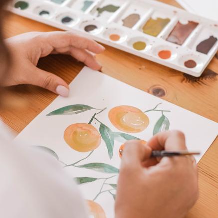 A woman is painting a picture of oranges and has a tray of colored paints in front of her.