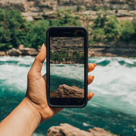 A hand holds an iphone in front of a stream and a mountain from arm's length.