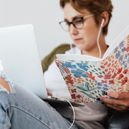 A woman wearing headphones looks at her laptop and reads a book at the same time.