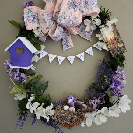 Image of a grapevine wreath with pink ribbon and purple and white flowers.