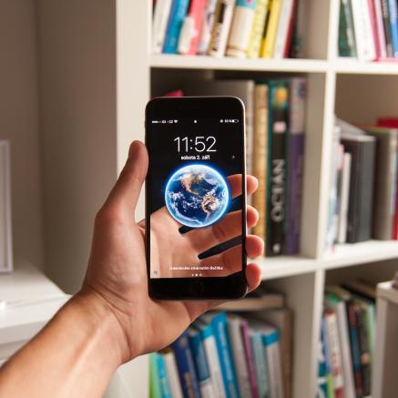 an outstretched arm holds an iphone in its hand in front of a book shelf.