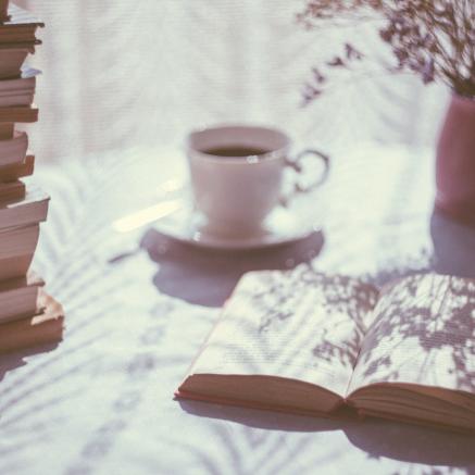 An open book sits on a table, beside a teacup and plant.