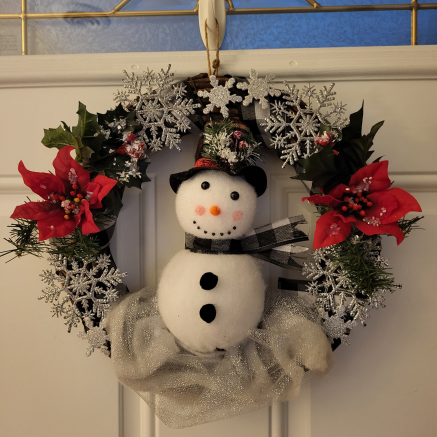 A snowman in the center of a holiday-themed wreath with poinsettias and snowflakes.