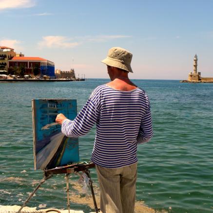 A man in a hat and a blue shirt paints the ocean while standing in front of the ocean.