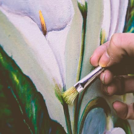 A hand holds a paintbrush on a painting of a white flower with large green leaves.