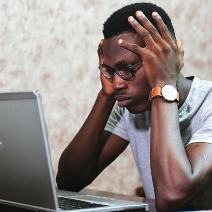 a young african-american man holds his head in frustration in front of his laptop.