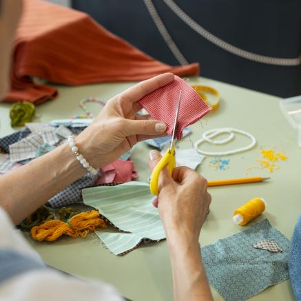 Image of woman's hands holding scissors and fabric, over a messy table with crafting supplies