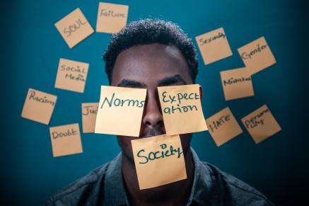 An African-American man stands with sticky notes on his face and the background. The notes on his face read "Norms, Expectations, Society"
