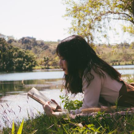 A young woman sits by the water, reading a book.