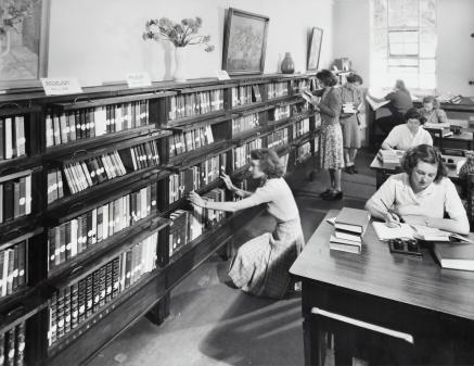 Black and white photo of women studying in a library, and browsing the shelf.