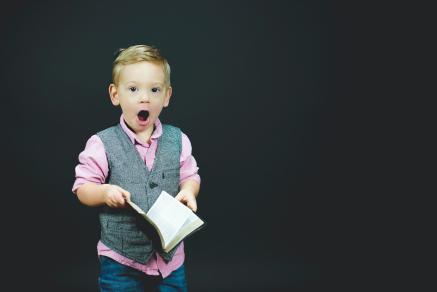 A boy in a vest holding a book and looking surprised.
