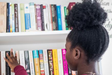 A Black girl looks at books on a library shelf.