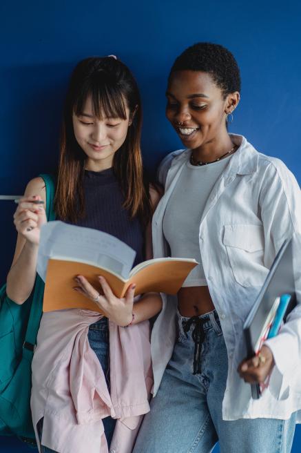 Two young women (one Asian and the other Black) leaning against a blue wall studying a book