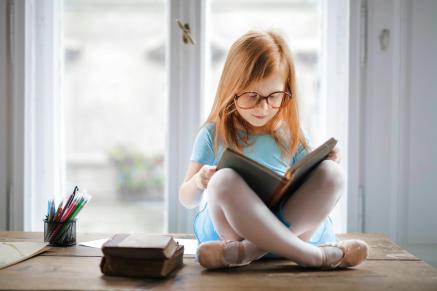 A young girl sits by a window reading a book.