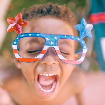 A young child shouts in celebration while wearing glasses with the American flag design on them