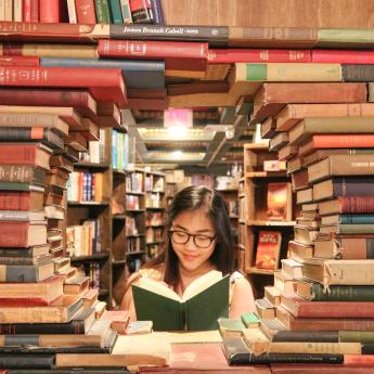 Young woman reading a book with a green cover, surrounded by books.