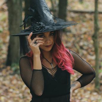 A woman with a pointy hat and pink hair stands in the woods.