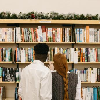 The back of a man and a woman facing a library bookshelf with an evergreen garland strand on top