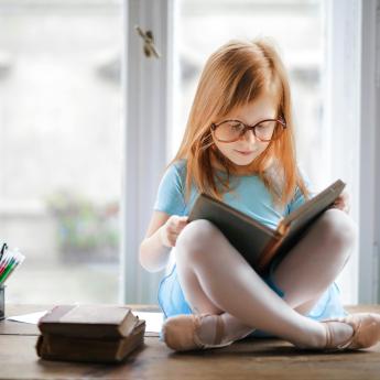 A young girl sits by a window reading a book.
